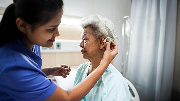 Nurse fitting a hearing aid in an elderly woman's ear in a hospital room.